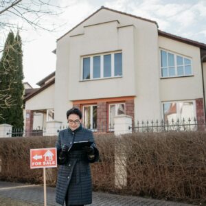 A real estate agent standing outside a house with a 'For Sale' sign, ready for viewing.