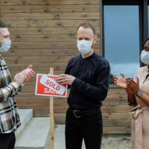 Real estate agent and clients celebrate sale with masks and a sold sign outdoors.